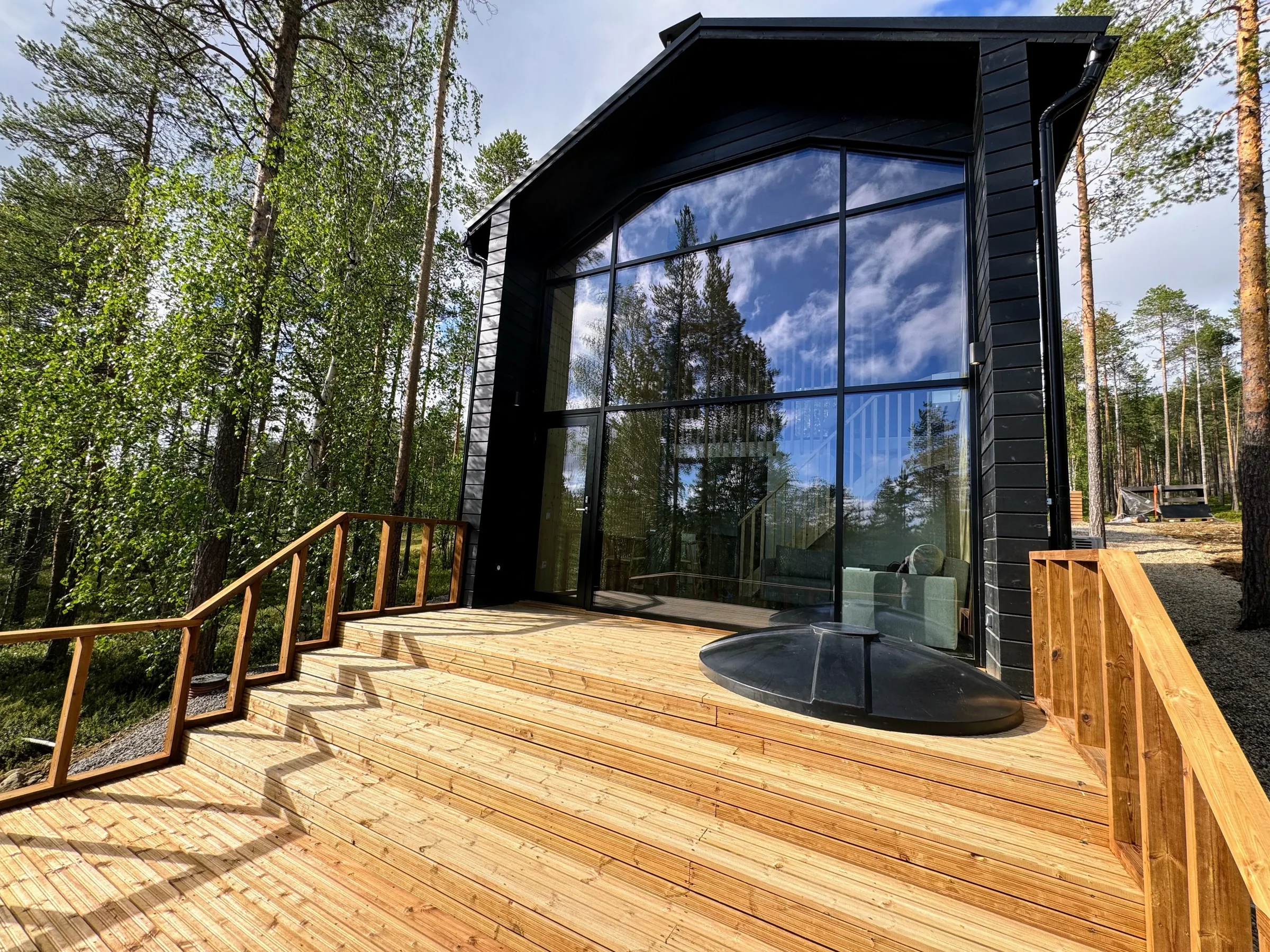 A black timber house with a glass facade and sunlit deck among summer trees