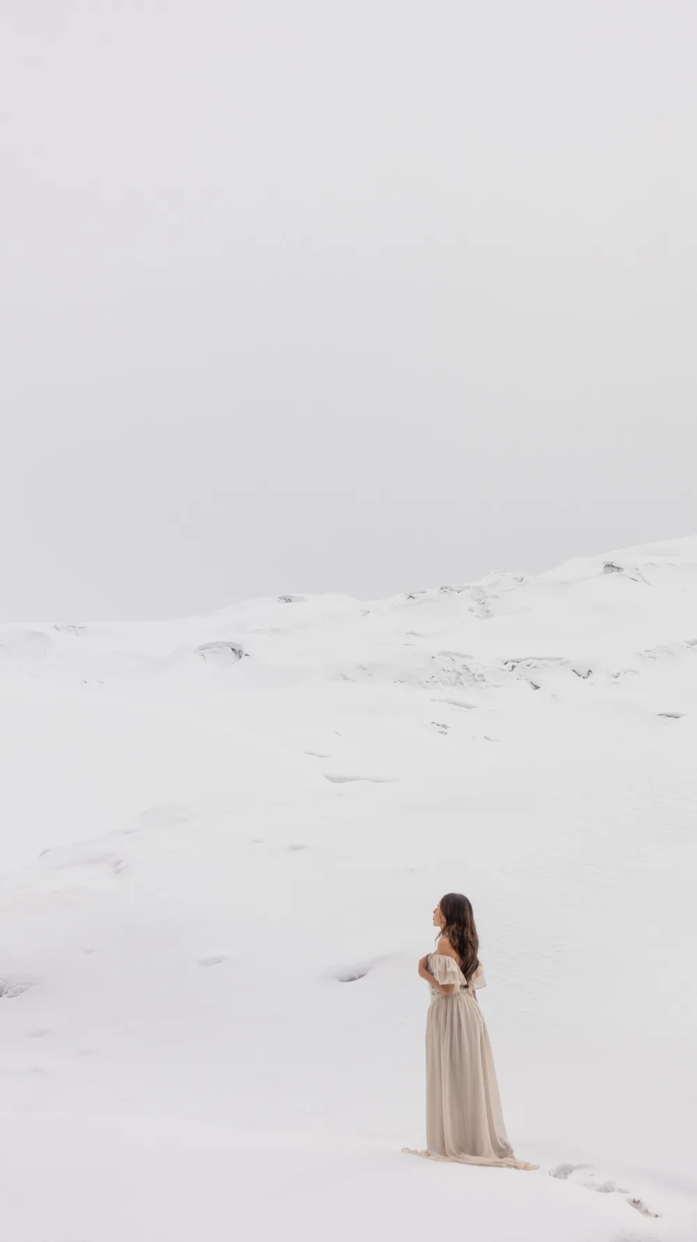 A woman in a pale ivory dress standing small in the lower third of a vast white snowfield